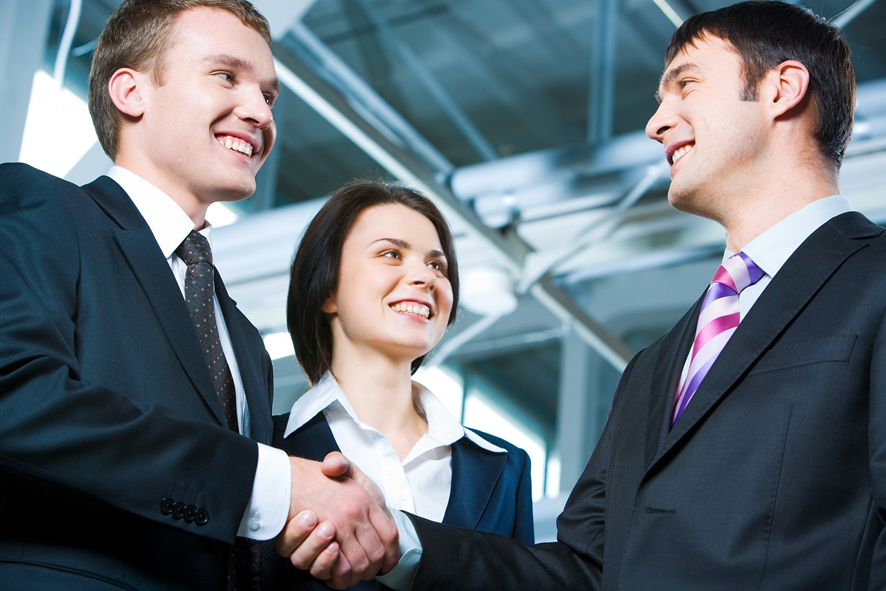 2 homens e uma mulher vestidos com ternos escuros num ambiente de trabalho se cumprimentando e sorrindo.